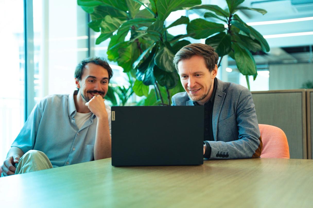 Zwei lächelnde Männer blicken in einem Büro auf einen Laptop.