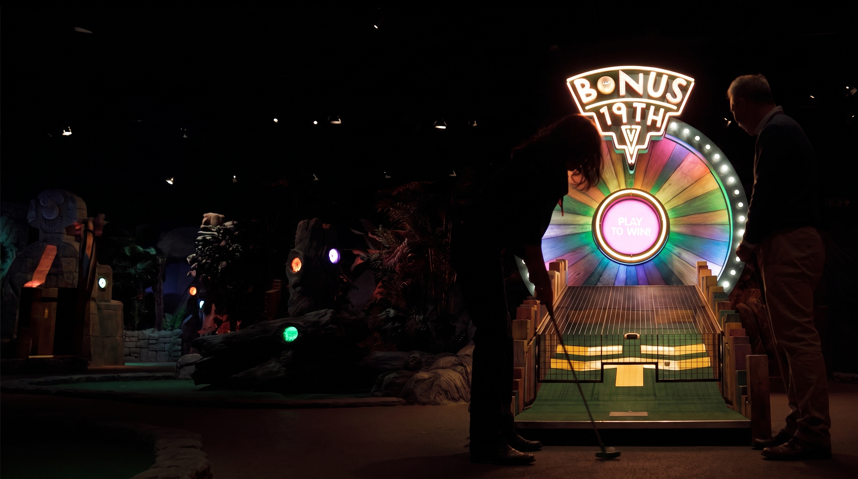 Zwei Personen spielen Minigolf auf einer dunklen, neonbeleuchteten Bahn und putten auf ein großes, buntes „Bonus 19th“-Rad.