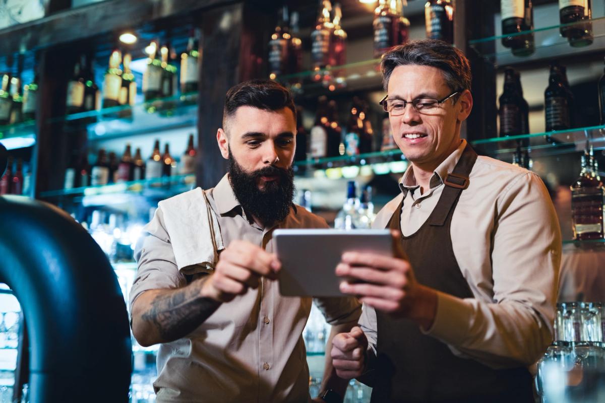 Two bar staff in aprons looking at a tablet together behind the bar
