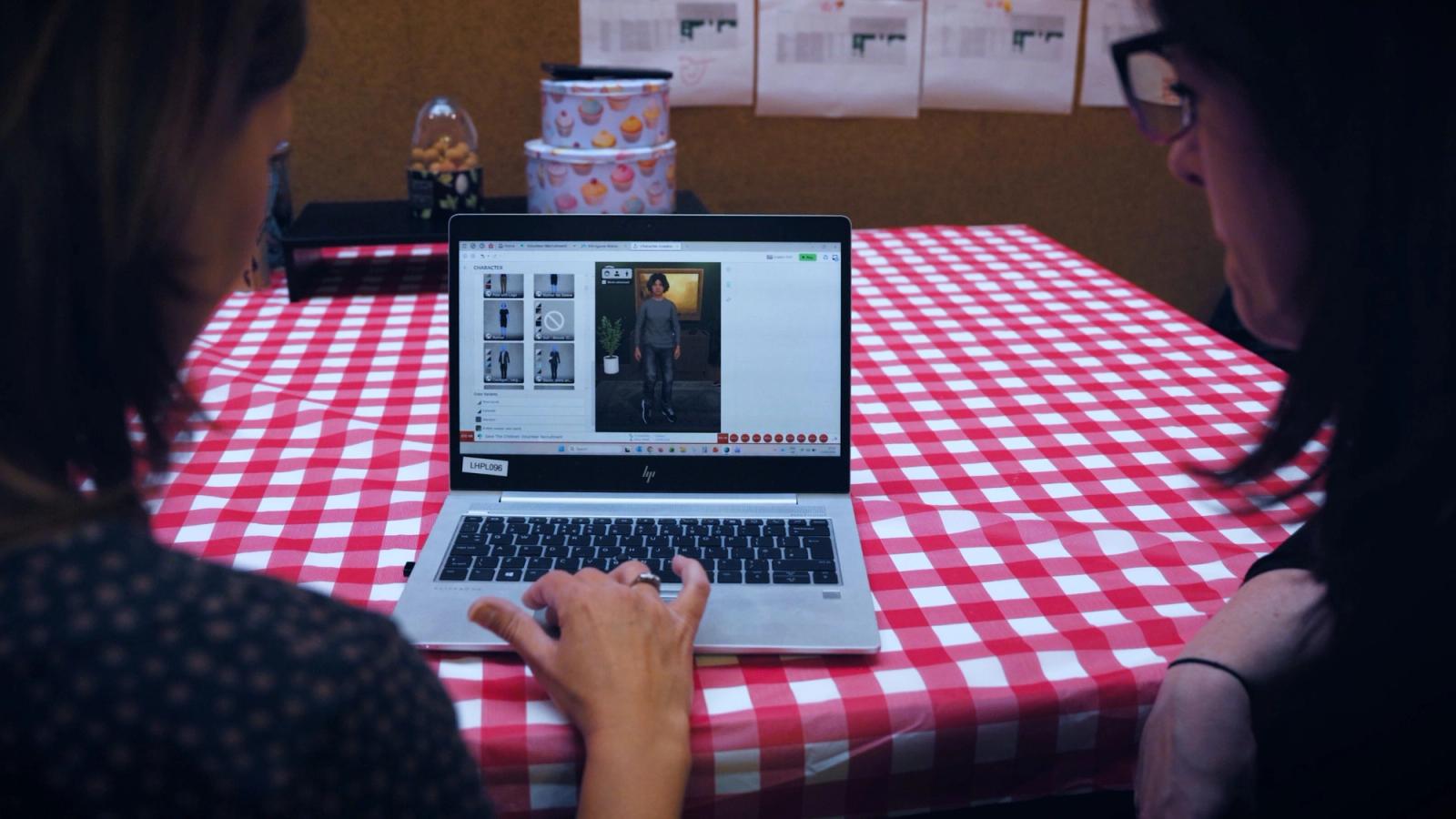 Zwei Personen an einem rot karierten Tisch schauen auf einen Laptop, der eine Figur in einem Avatar-Editor zeigt
