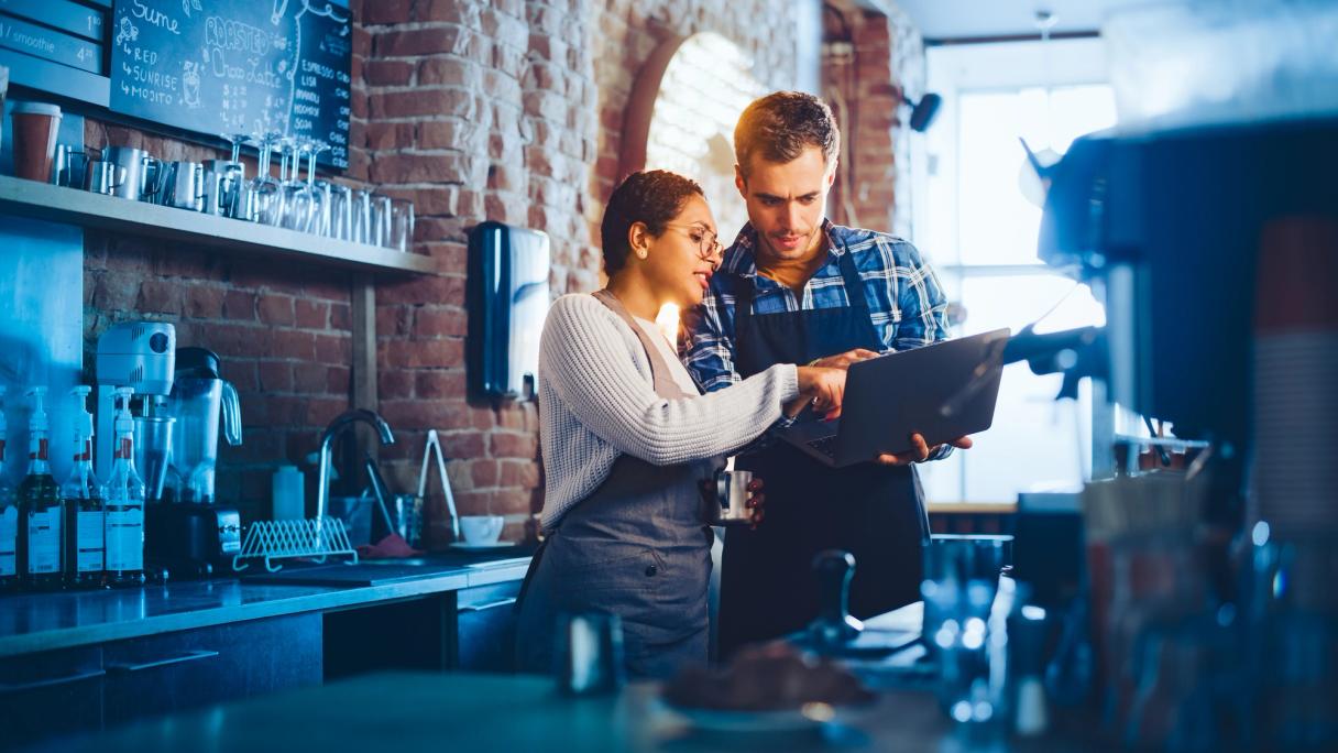 Zwei Café-Mitarbeitende in Schürzen schauen hinter der Kaffeebar auf einen Laptop.