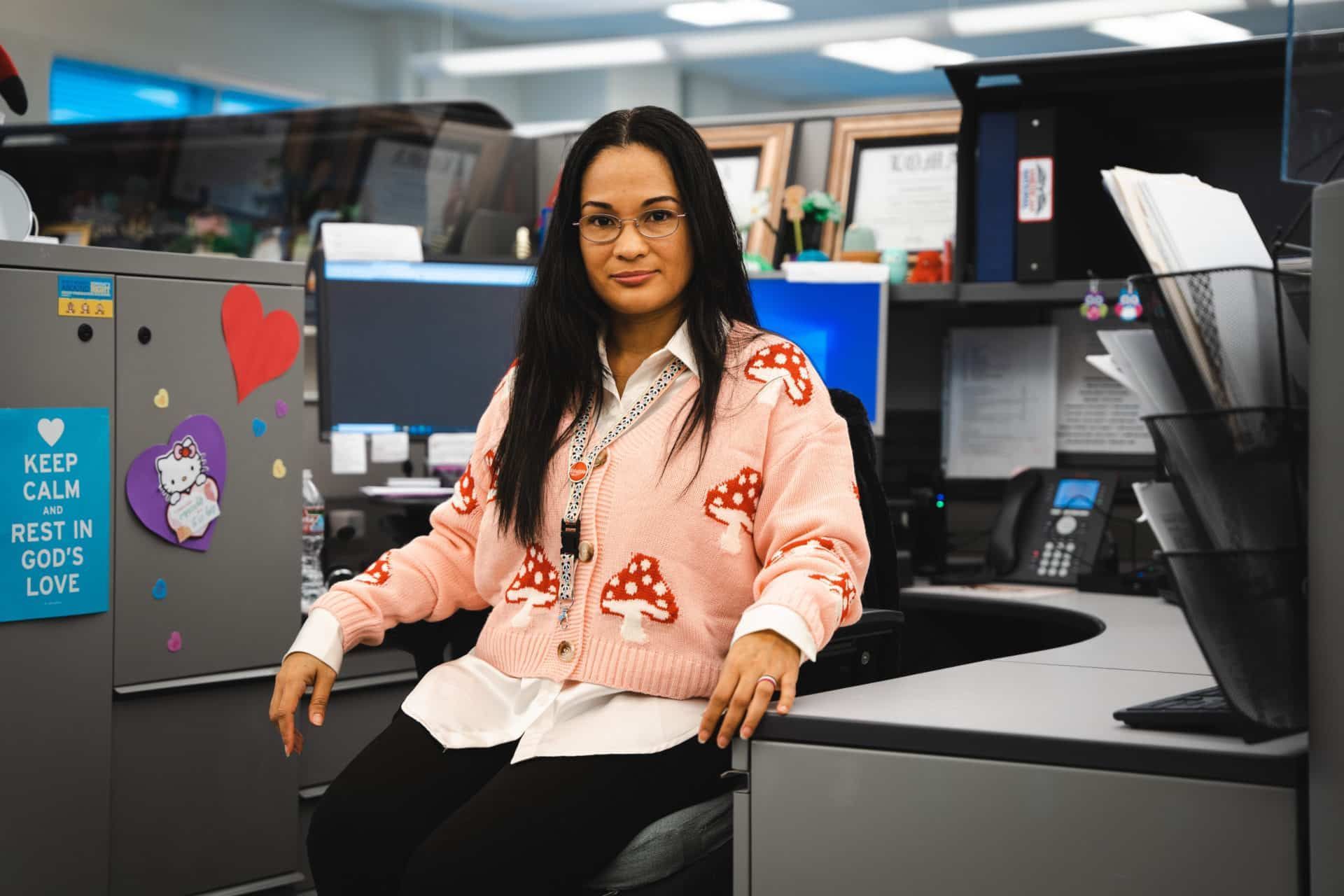 Claudia Rosencranz sitzt auf einem Bürostuhl in ihrer Arbeitsnische und trägt eine rosa Strickjacke mit Pilzmuster