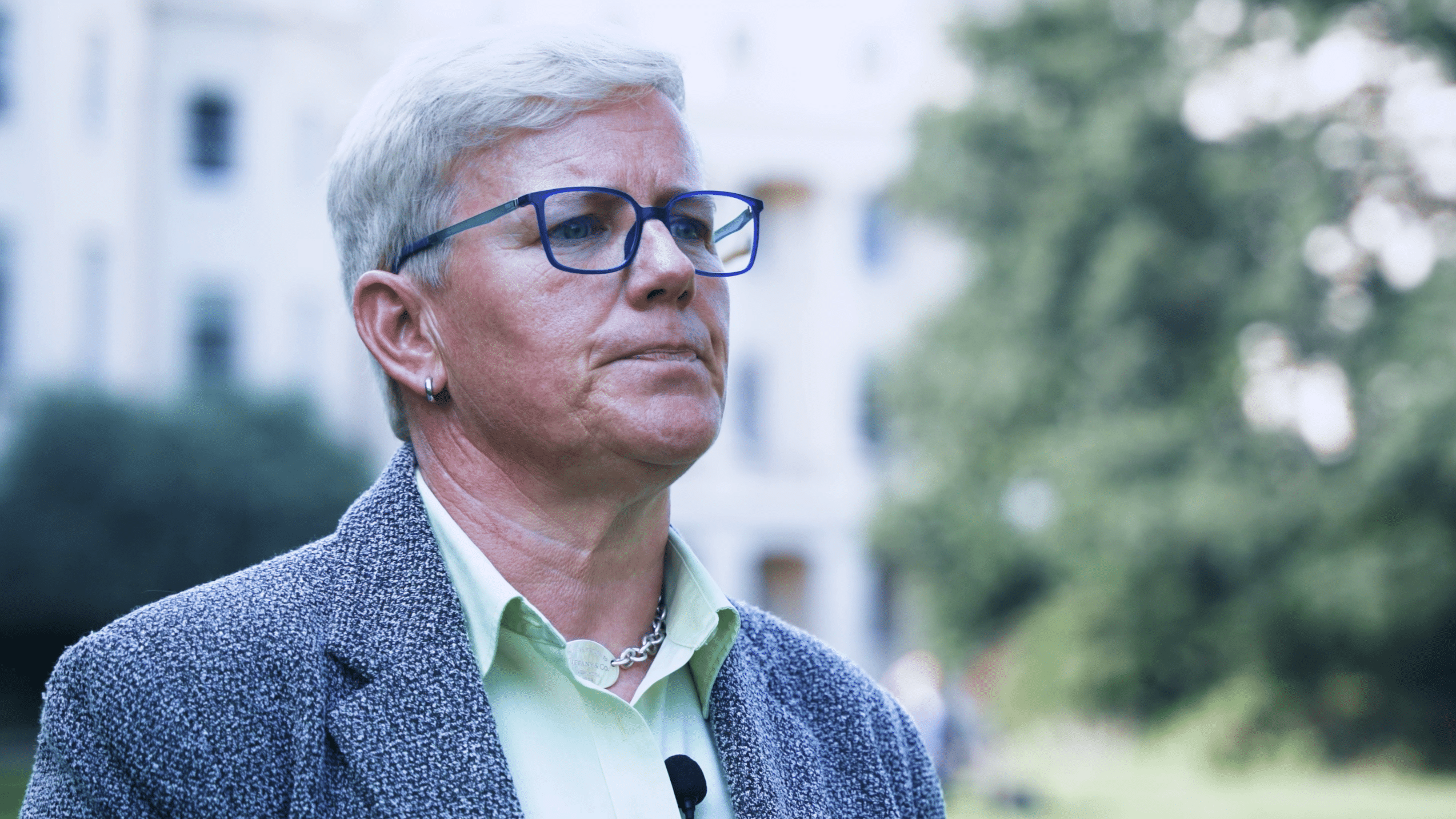 Michele Asbury in grauem Blazer und mit blauer Brille, im Freien vor einem weißen Gebäude