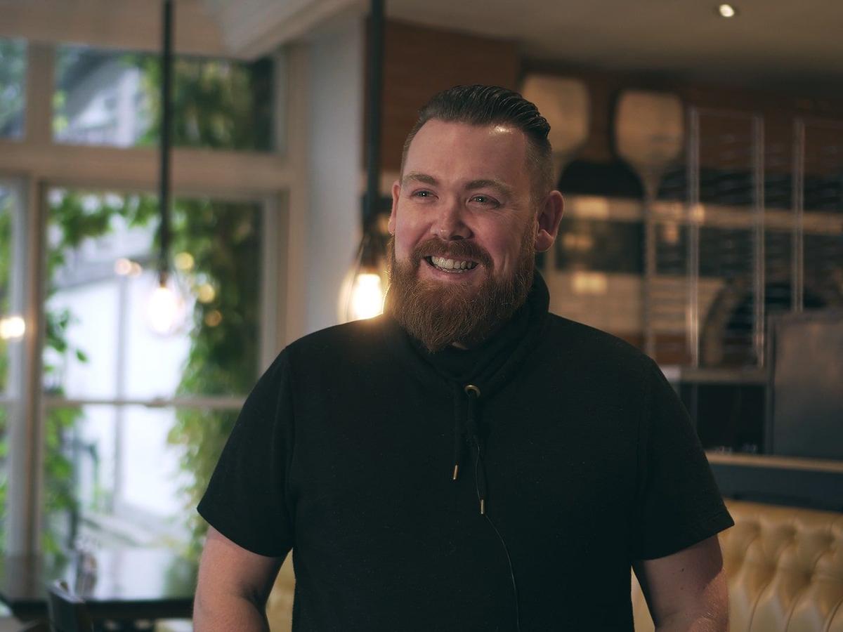 Lächelnder Mann mit Bart in schwarzem Hemd in einem Pub mit Hängeleuchten und einer Lederbank