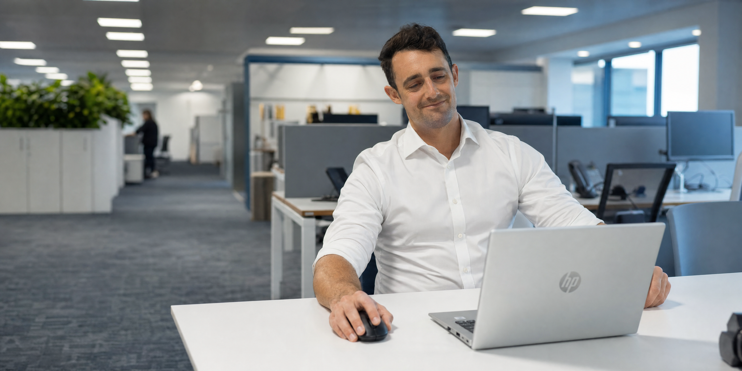 A man with a slight smile works on a laptop at a desk in a modern office.