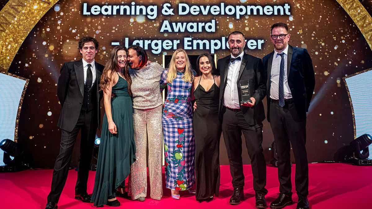 Sieben Personen auf einer Bühne, eine hält eine gläserne Trophäe, unter einer Leinwand mit der Aufschrift „Learning & Development Award – Larger Employer“