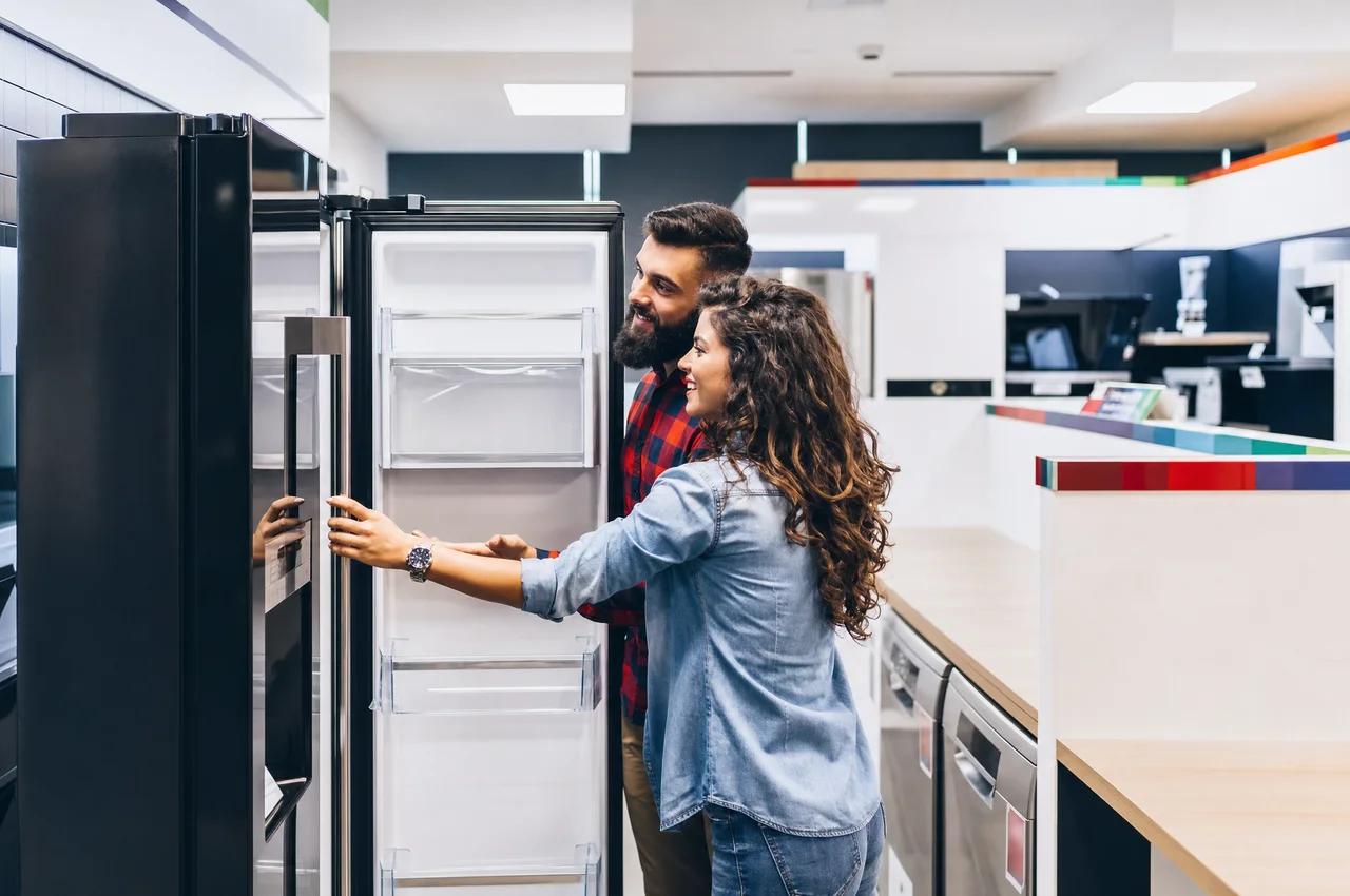 A smiling couple looks inside an open black refrigerator at an appliance store.