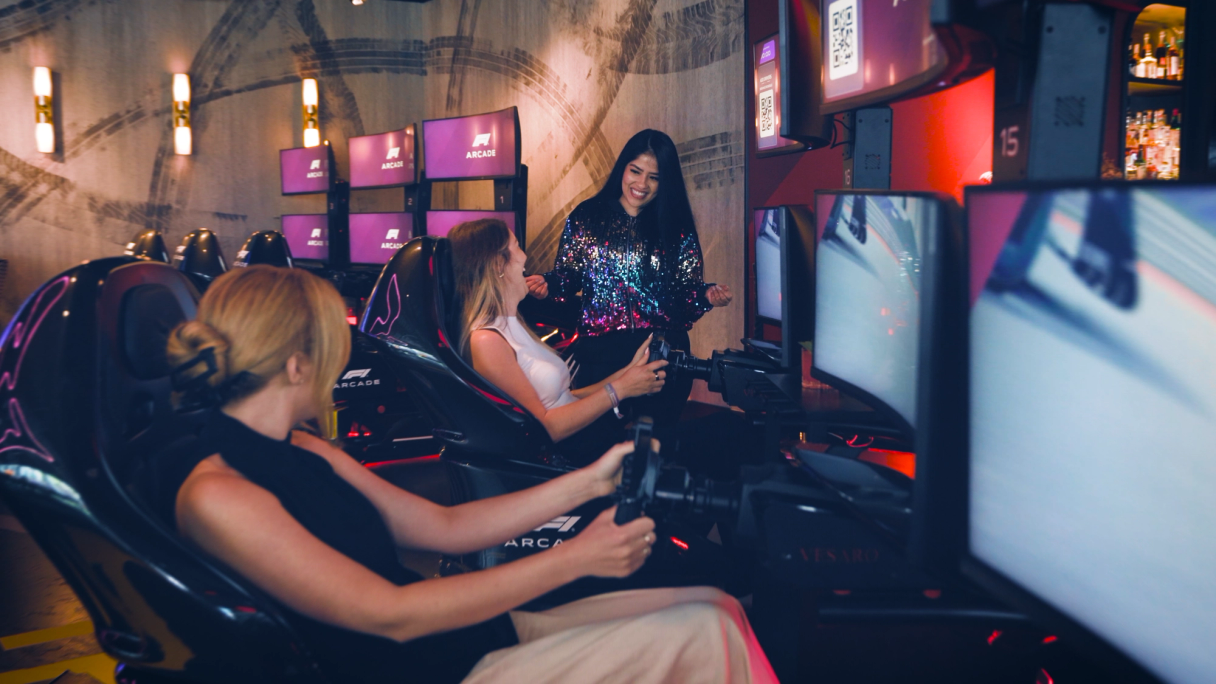 Zwei lachende Personen in Rennsimulator-Sitzen, eine dritte steht daneben, im F1 Arcade