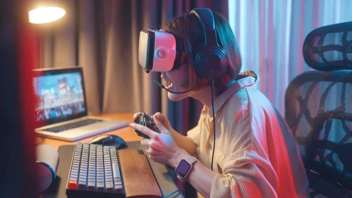 Person med VR-headset och hörlurar håller en spelkontroll vid ett skrivbord med laptop
