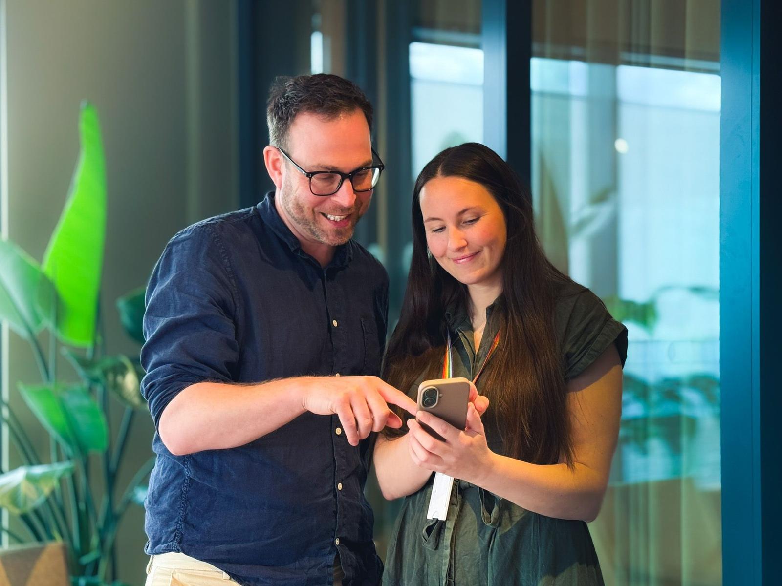 A man points at a phone screen held by a woman as the two look at it together indoors