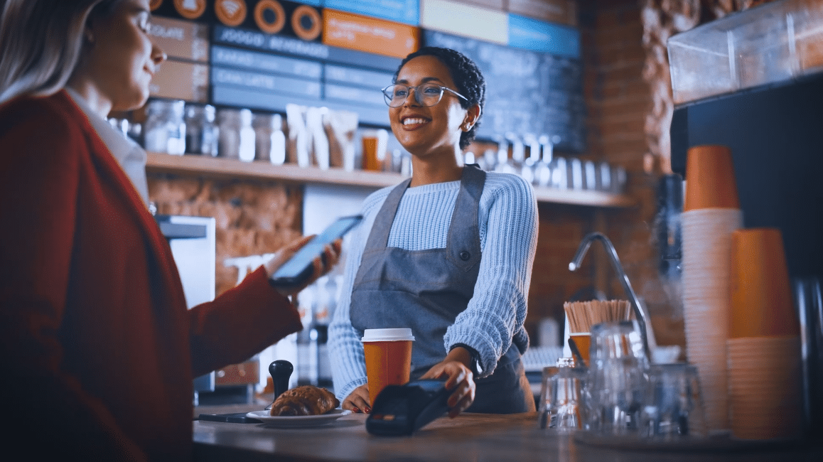 Lächelnde Barista in Schürze bedient einen Kunden, der am Café-Tresen per Smartphone bezahlt