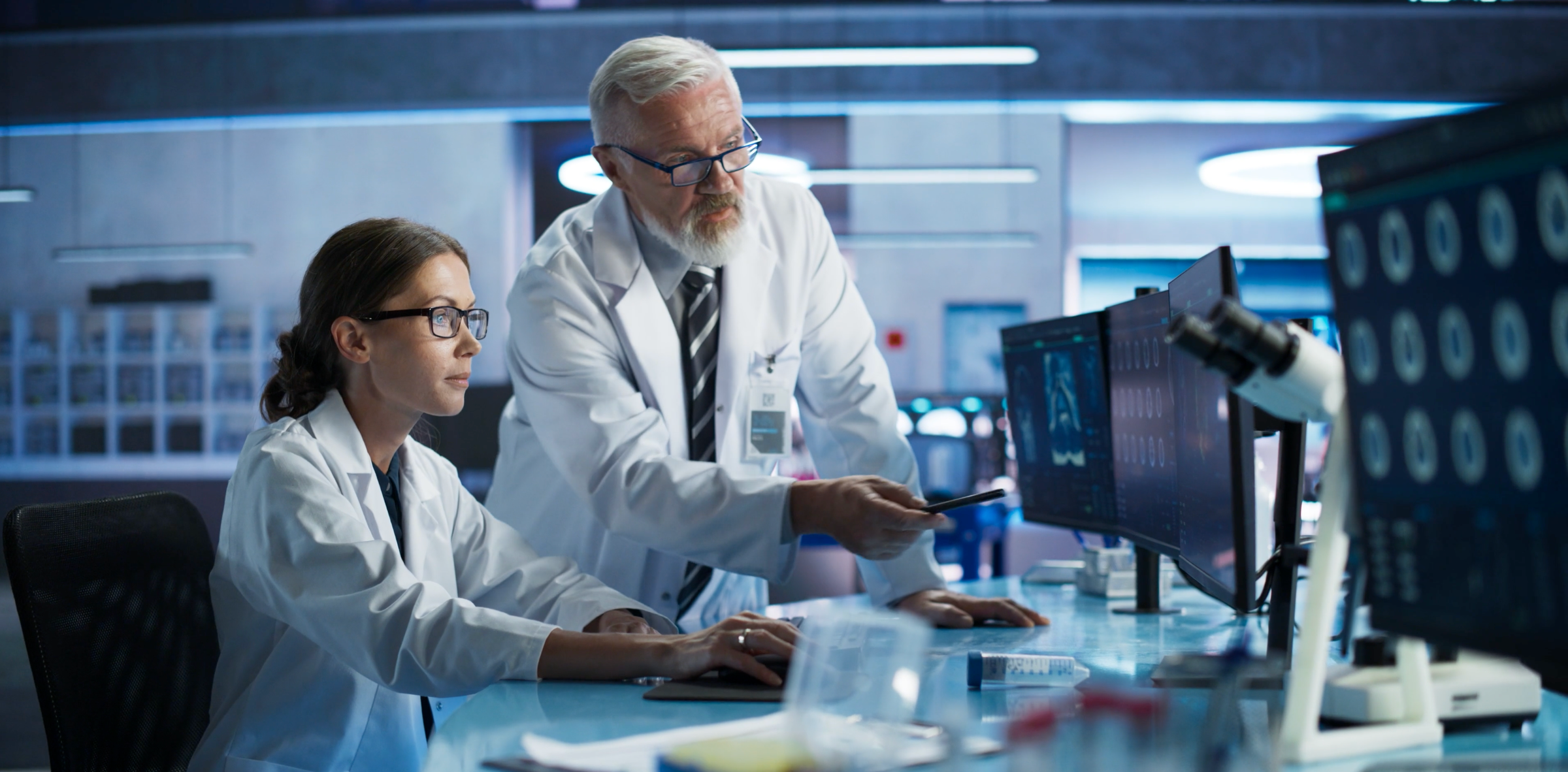 Two scientists in lab coats review medical scans on computer monitors in a laboratory.