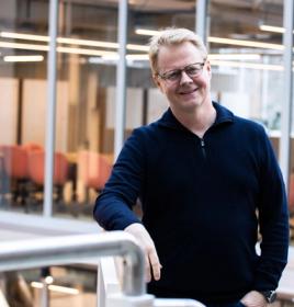 Trond Aas lächelt in einem dunklen Troyer an einem Geländer in einem Büro mit Glaswänden