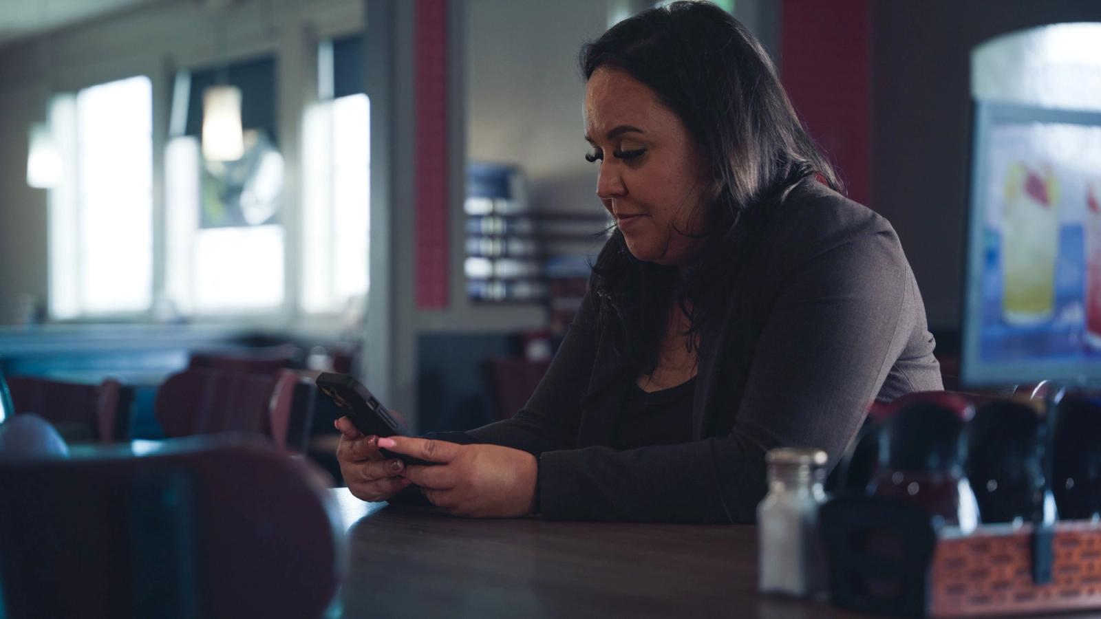 Eine Frau sitzt an einem Tisch in einem schwach beleuchteten Restaurant und schaut neben einem Gewürzständer auf ihr Smartphone
