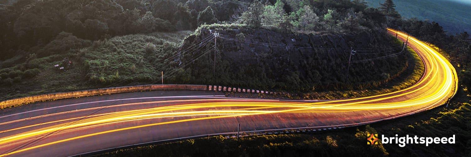 Traînées de lumière le long d'une route sinueuse au crépuscule, avec le logo Brightspeed