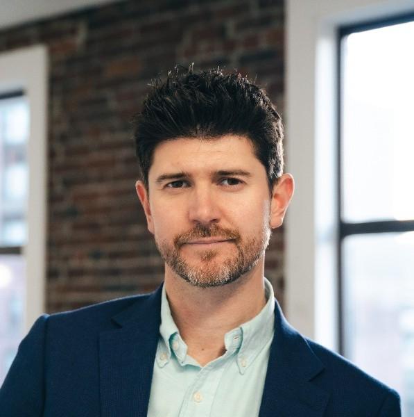 Man with dark hair and a short beard in a navy blazer, in a loft with exposed brick