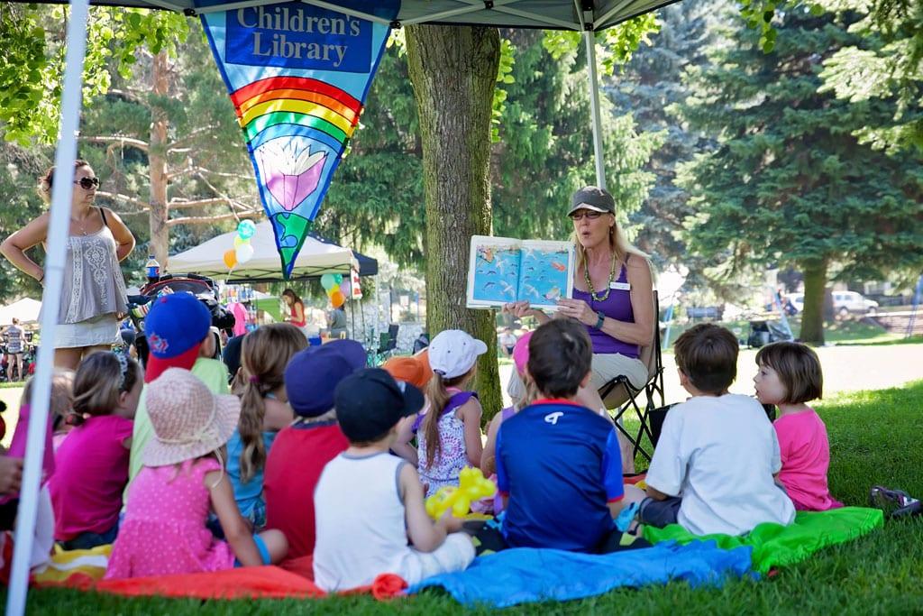 Frau liest einer Gruppe kleiner Kinder auf dem Rasen unter einem Zelt mit der Aufschrift „Children's Library“ aus einem Bilderbuch vor
