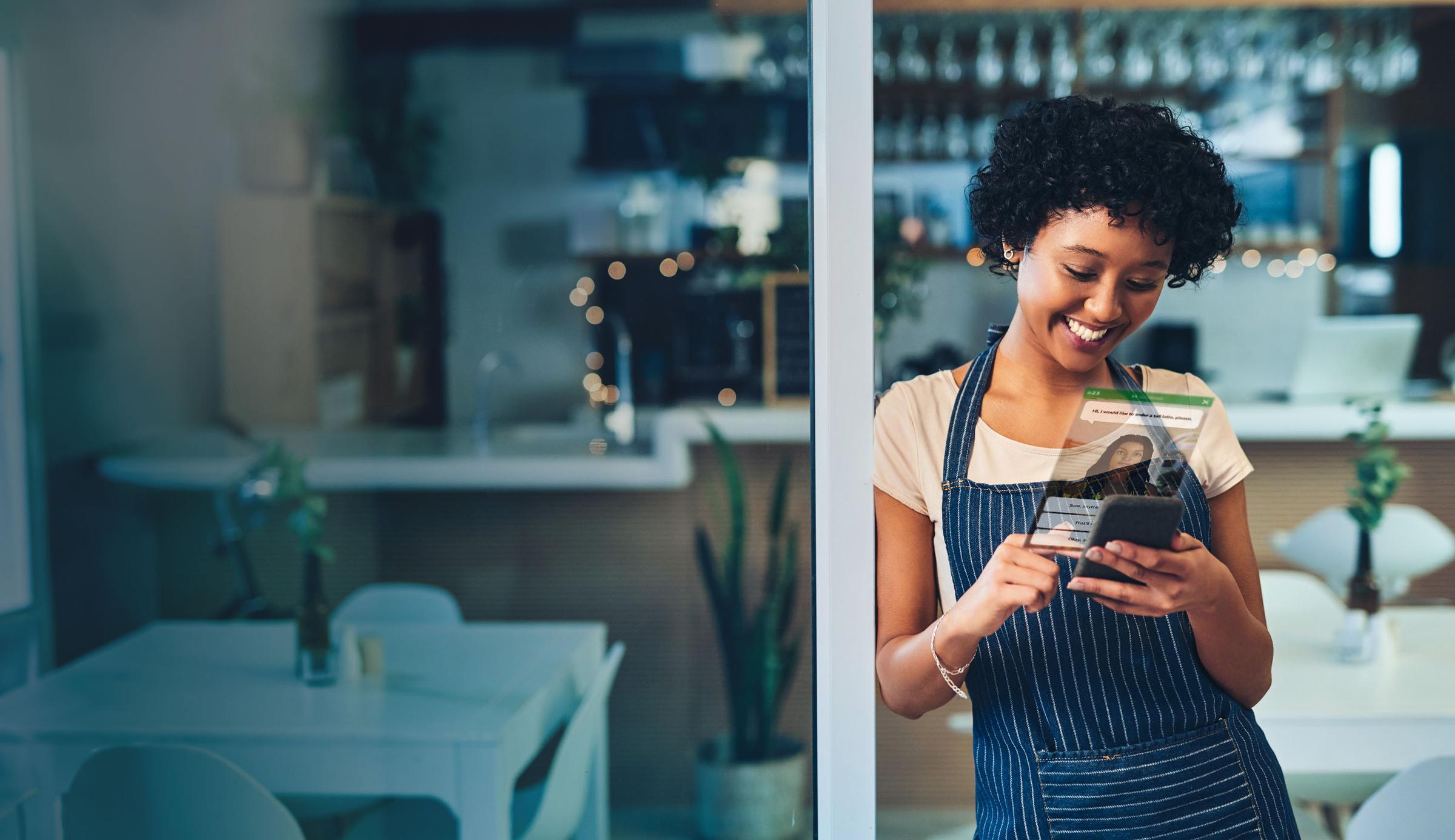 A smiling woman in an apron uses her smartphone with a digital overlay displaying a profile and data, in a café.