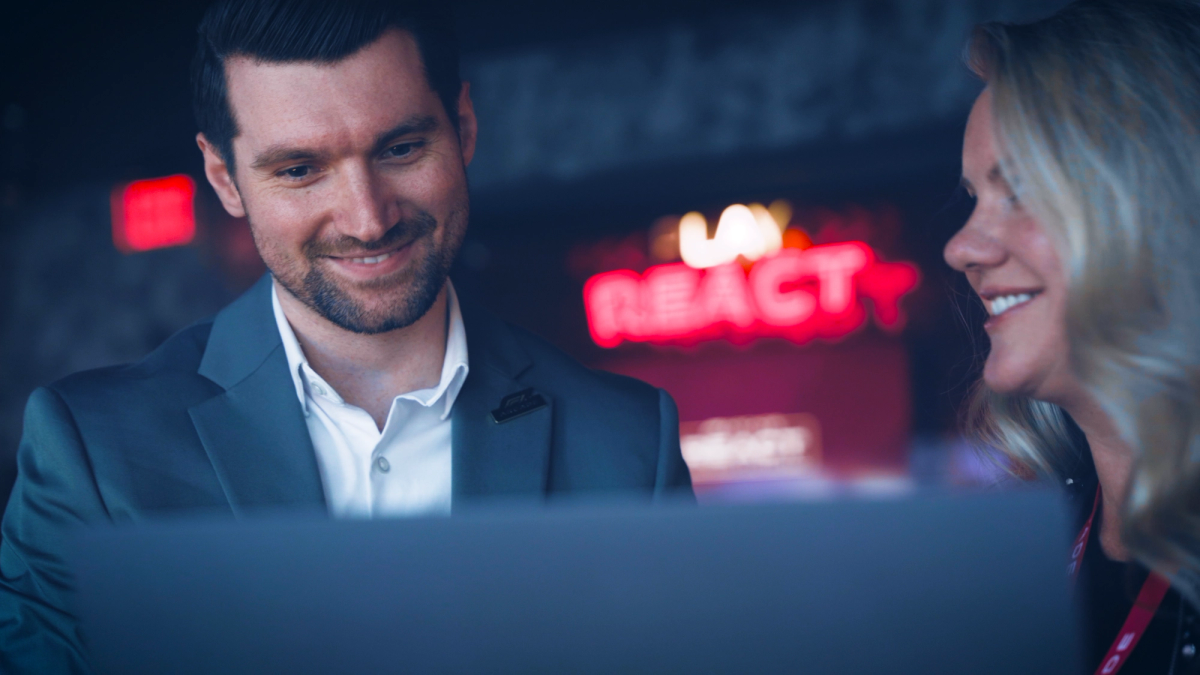 Mann und Frau lächeln vor einem aufgeklappten Laptop in einem dunklen Neonlicht-Ambiente