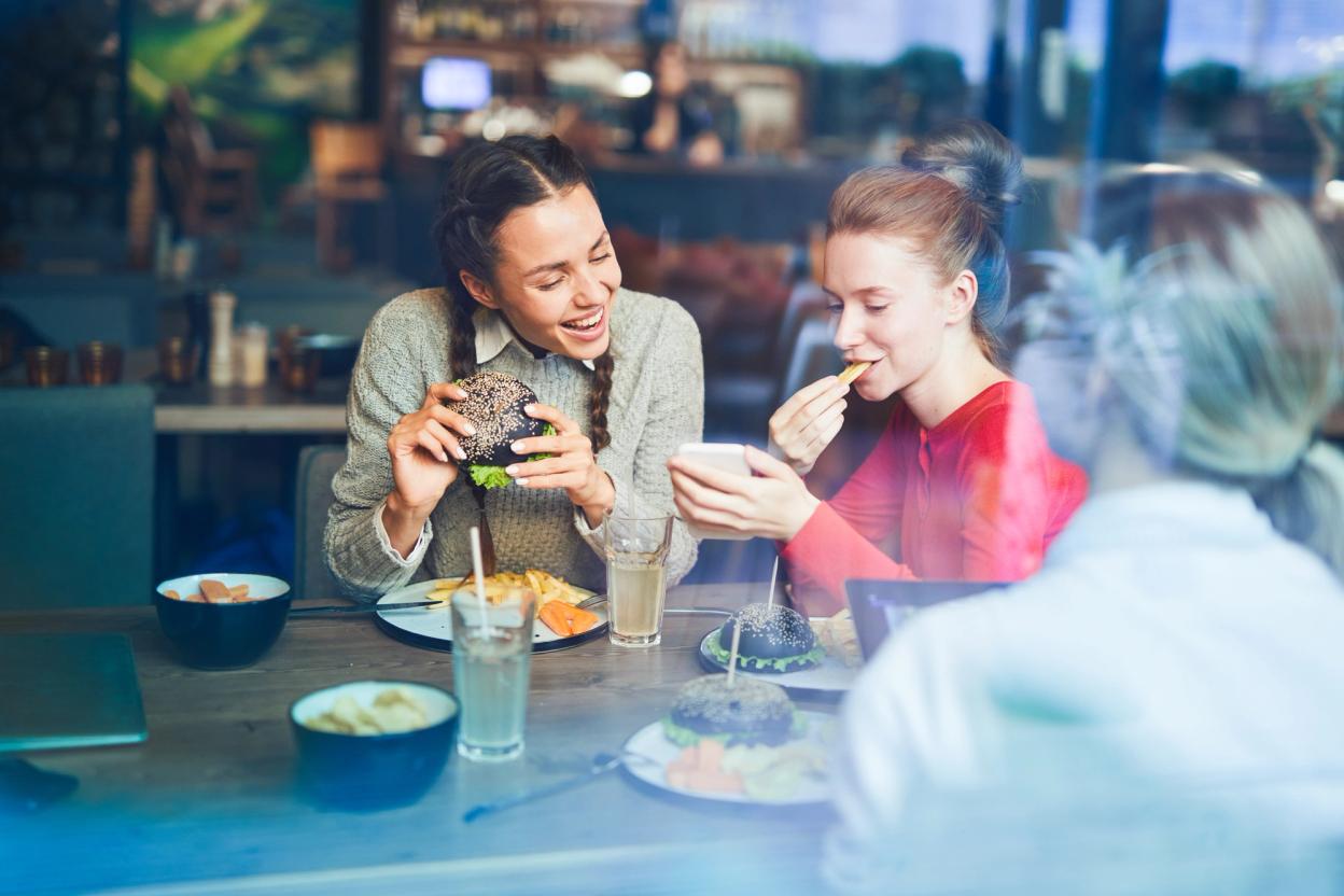 Zwei junge Frauen teilen sich Burger an einem Restauranttisch, im Vordergrund weitere Gäste
