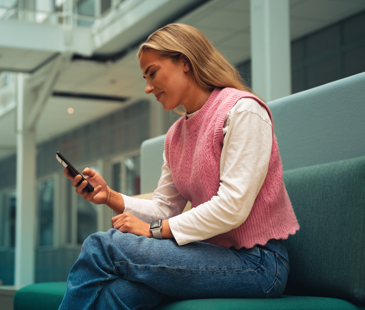 Junge Frau in pinker Strickweste sitzt auf einem grünen Sofa in einem Büro-Atrium und schaut auf ihr Smartphone