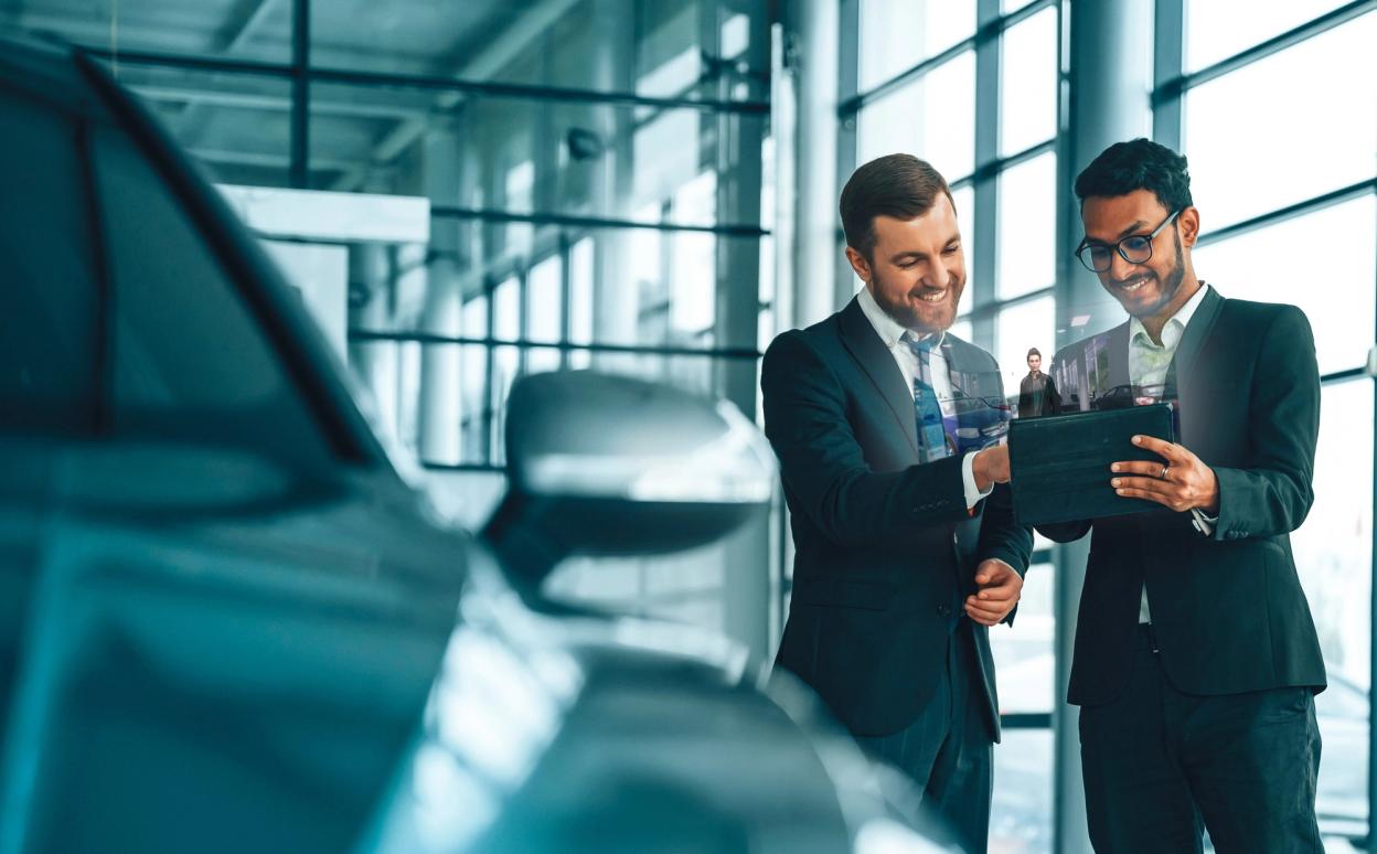 Autofabrik: Automobilingenieurin mit Schutzhelm steht und nutzt einen Laptop. Überwachung, Steuerung, Anlagenproduktion. Automatisierte Montagelinie mit Roboterarmen fertigt Elektrofahrzeuge.