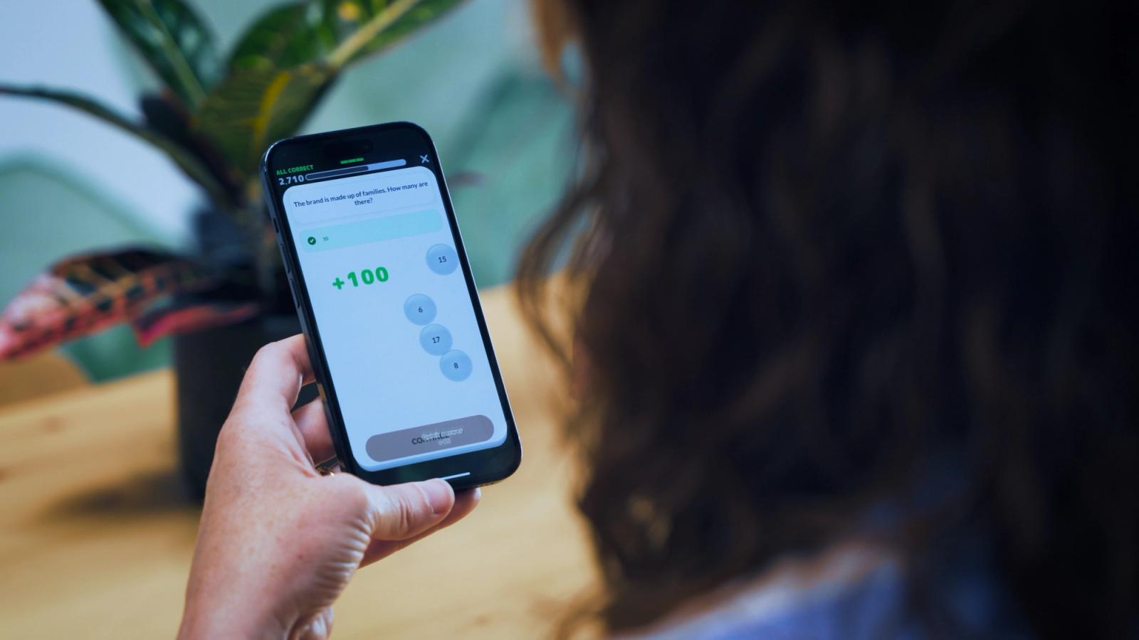 A hand holds a smartphone displaying a mobile app with "+100" on the screen.
