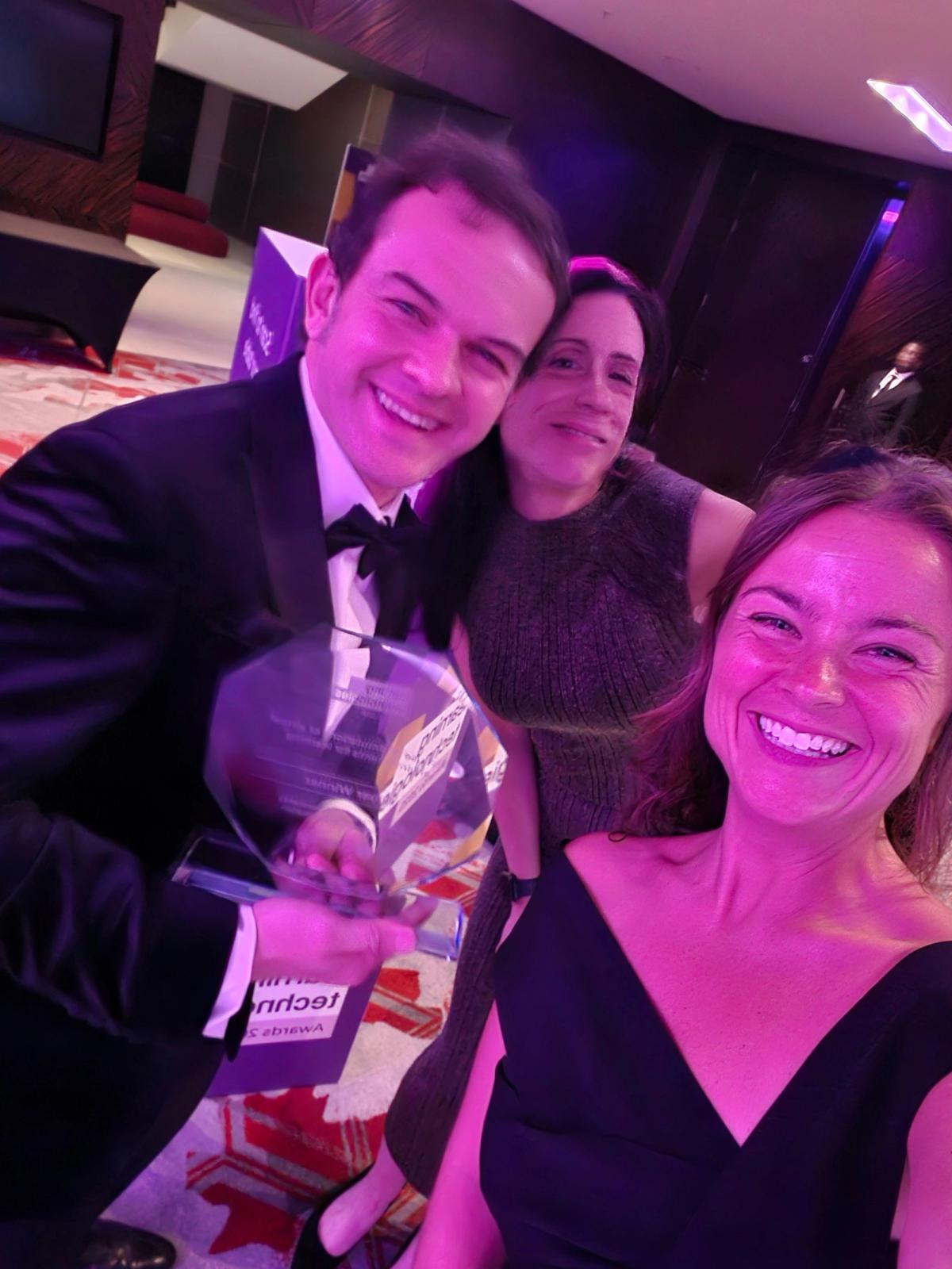 Close-up selfie of three people at an evening awards event, one in a tuxedo holding a glass trophy