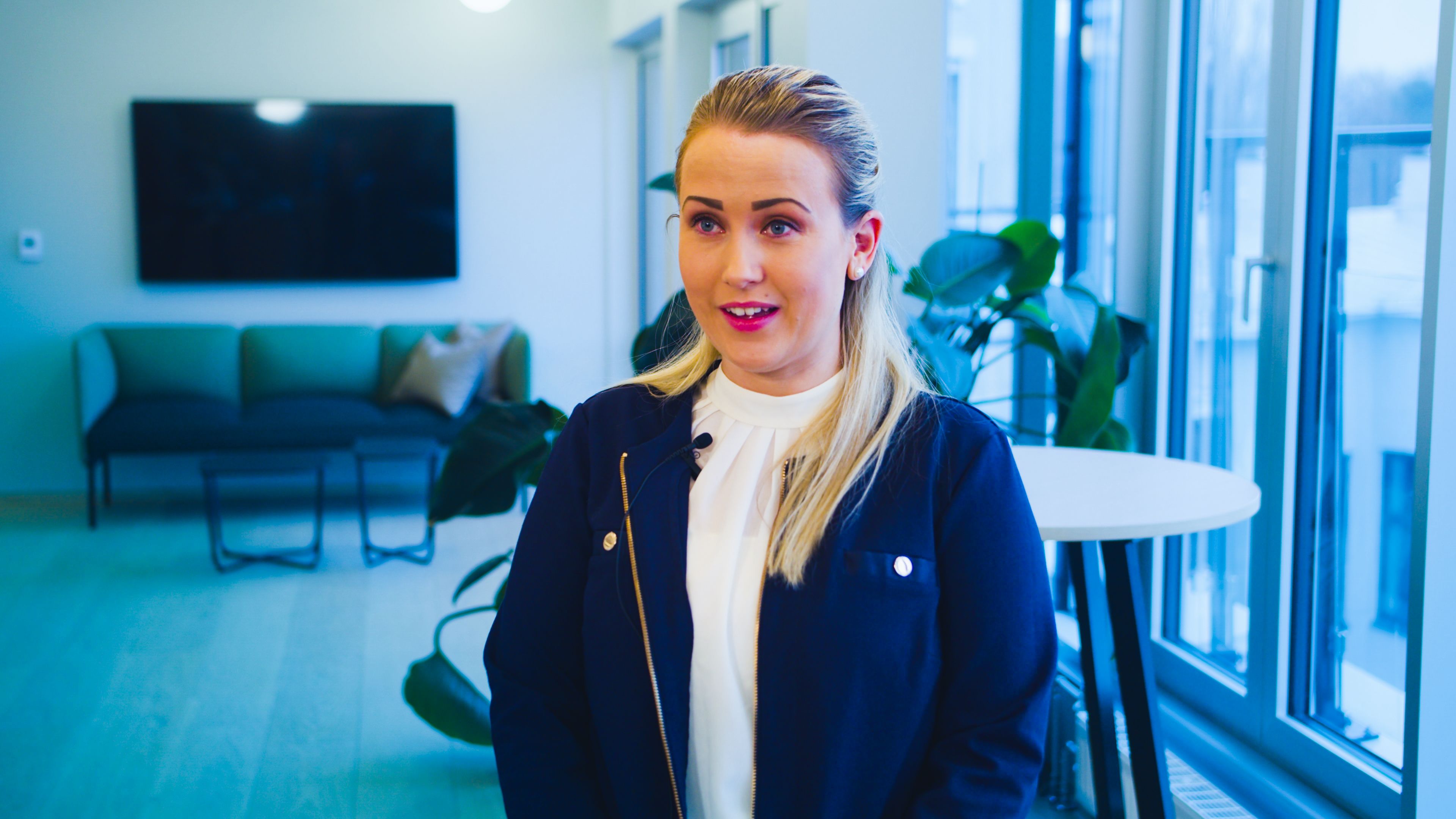 Victoria Barikmo steht in einer Büro-Lounge, hinter ihr ein Sofa und ein Bildschirm an der Wand