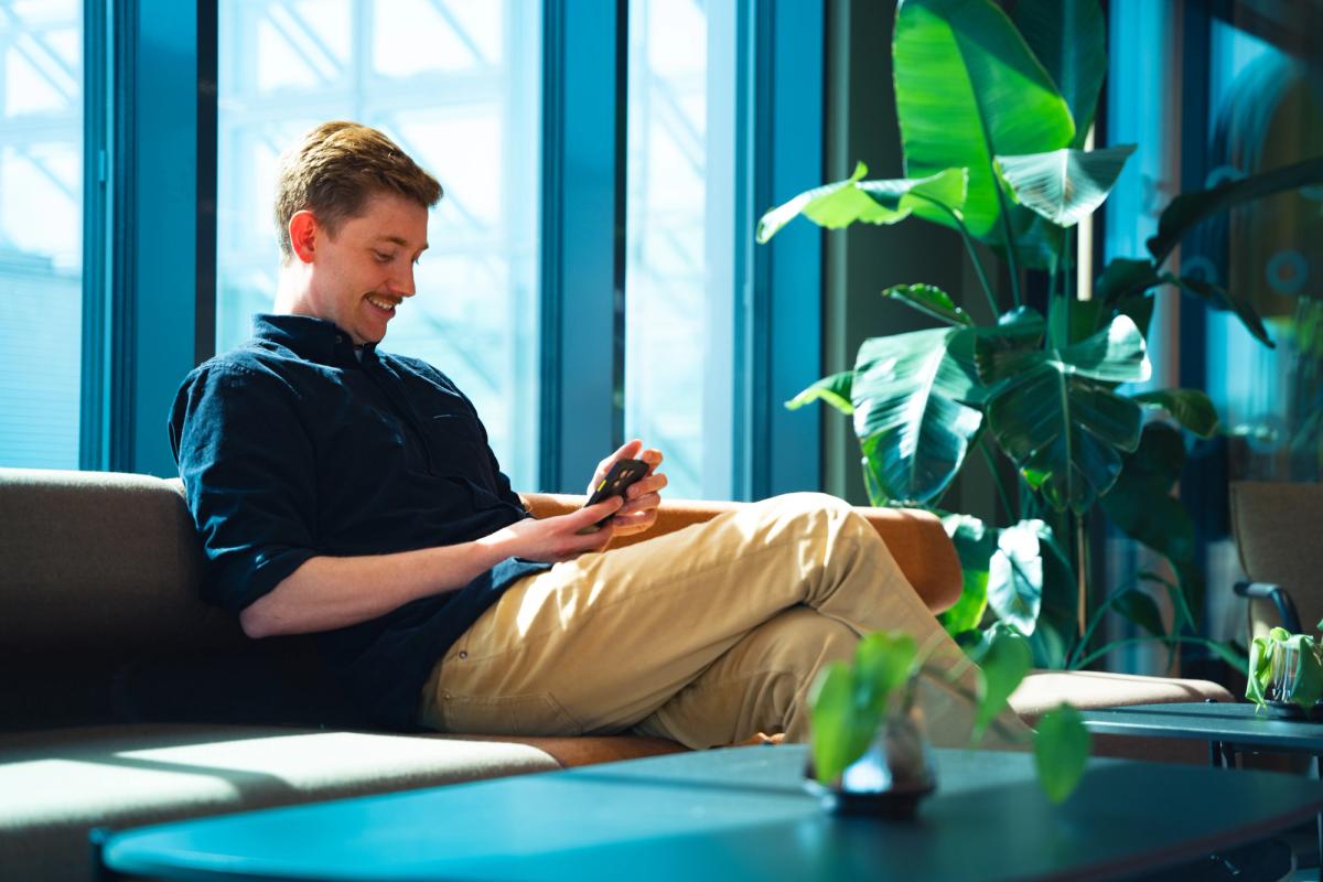 Mann lächelt auf sein Smartphone und sitzt auf einem Sofa vor hohen Bürofenstern