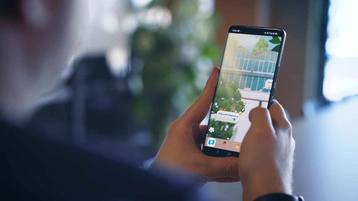 Hände halten ein Smartphone mit einem Handyspiel, das ein gezeichnetes Gewächshaus mit Blumen zeigt.