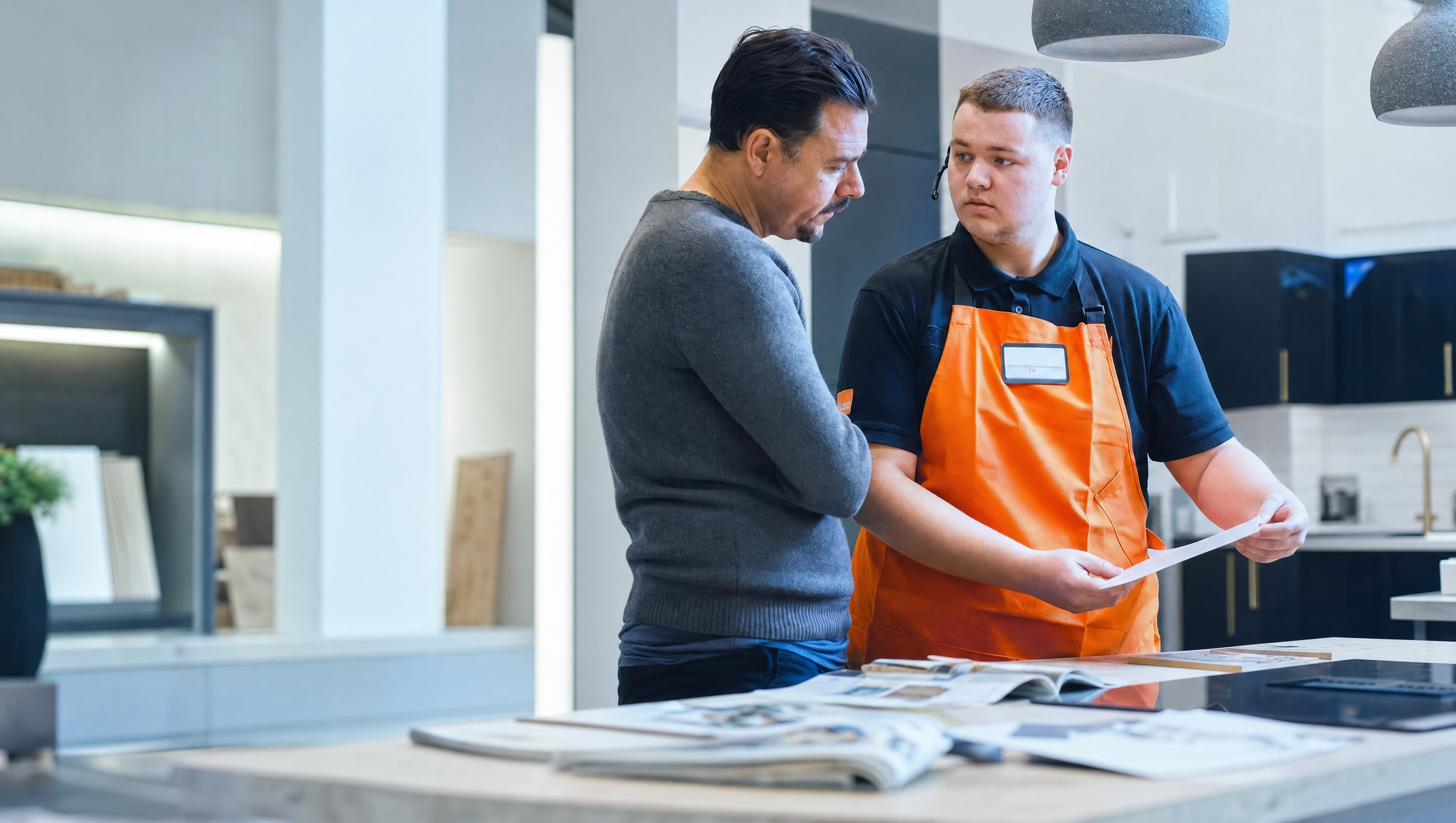 An employee in an orange apron discusses options with a customer in a home improvement showroom.