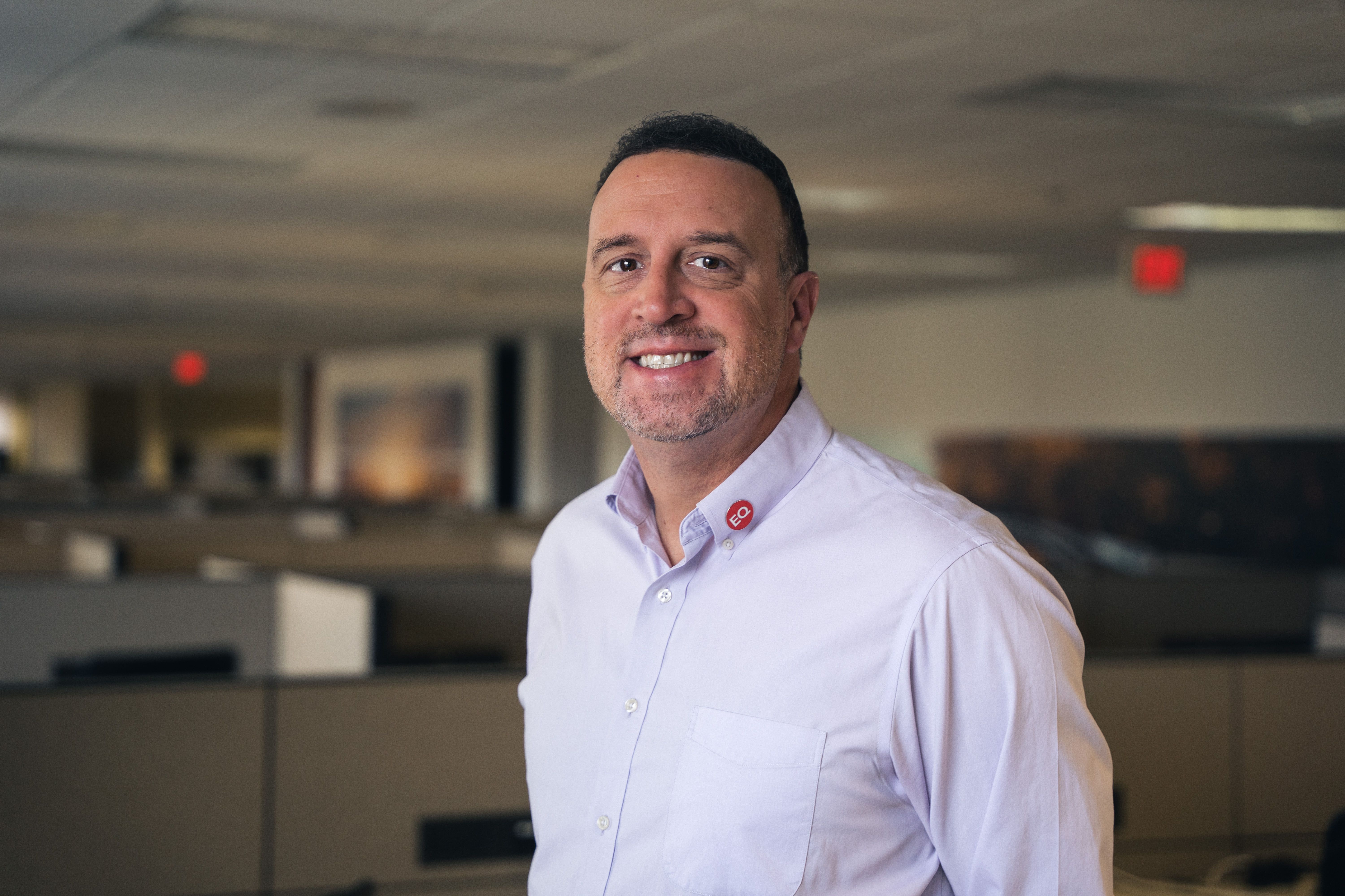John Horvath smiling in an office with cubicles behind him, in a light shirt with a small red pin
