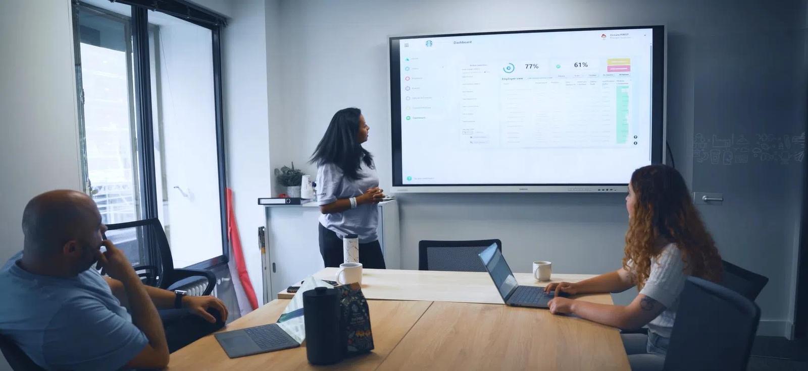 Three people in a modern meeting room, one woman presenting data on a large screen to two colleagues.