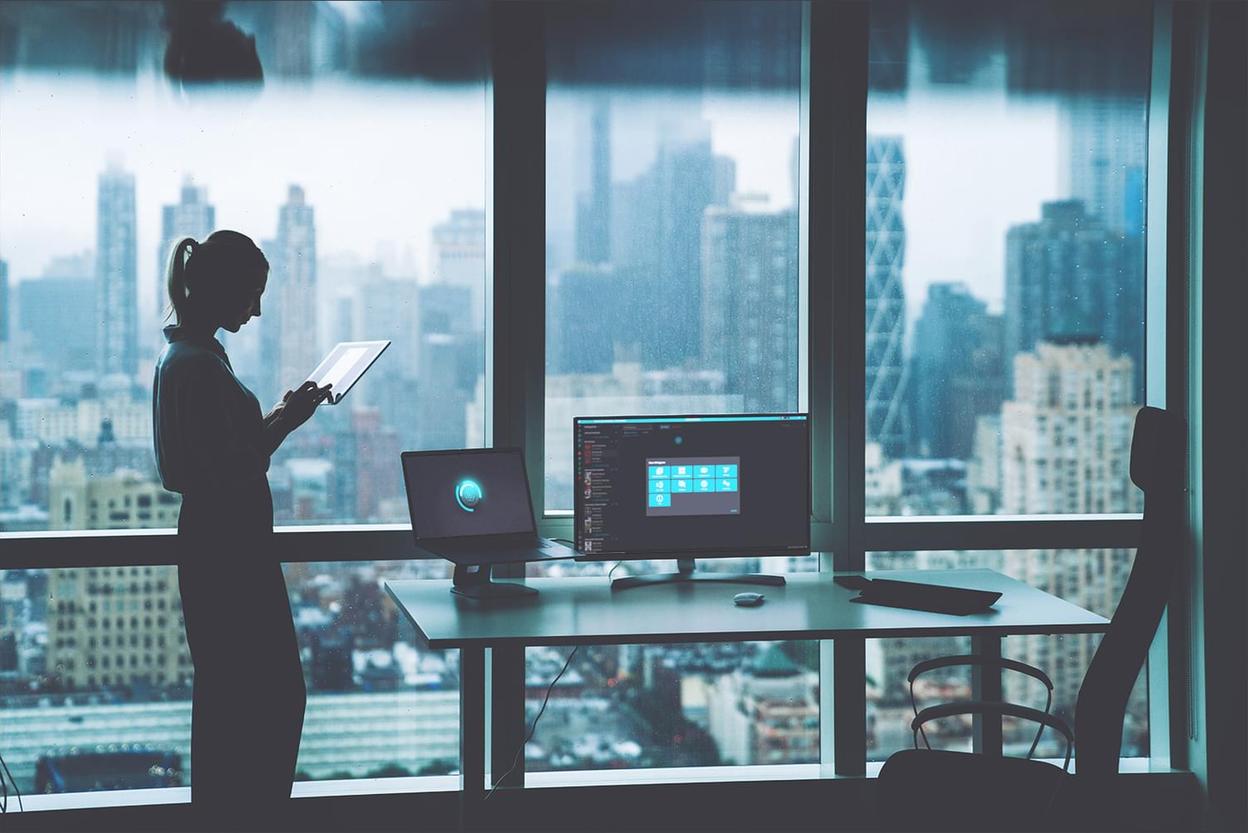 Frau mit Tablet am Bürofenster mit Blick auf die Skyline, Laptop und Monitor auf dem Schreibtisch