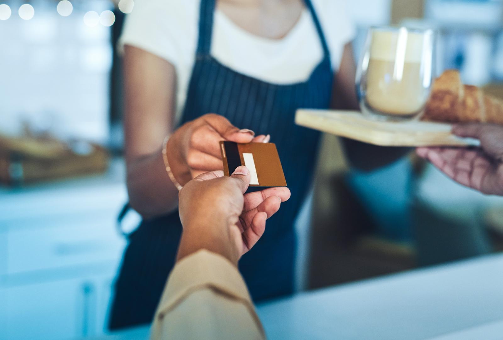 Two hands exchanging a card over a counter, with a latte and a croissant on a wooden board alongside