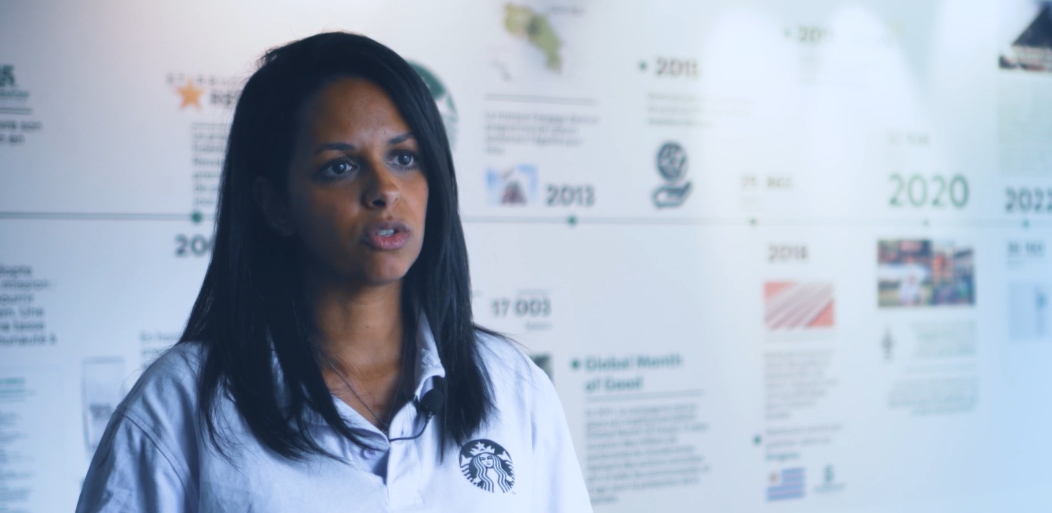 Woman in a Starbucks-branded polo shirt speaking in front of a company history timeline wall