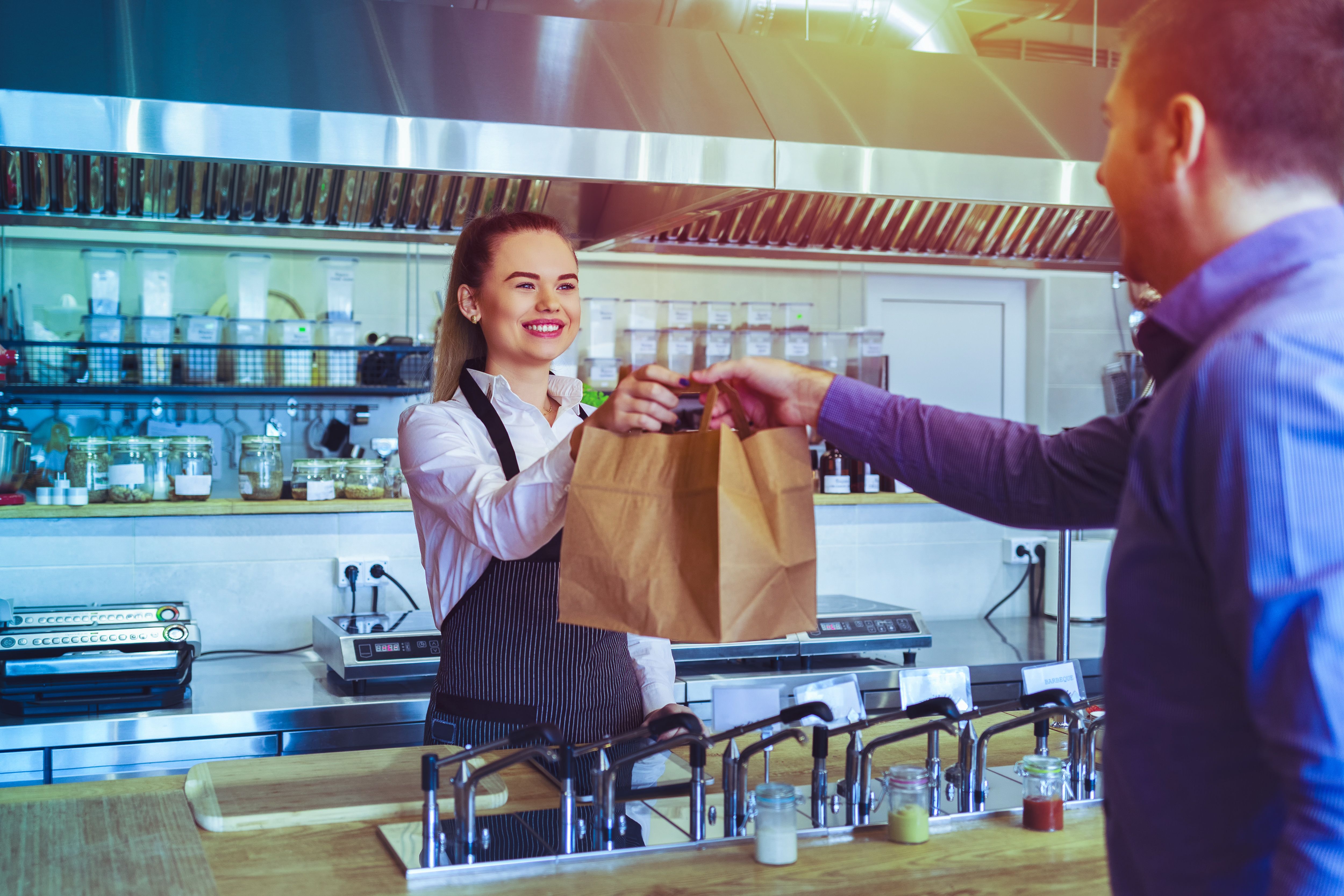 A smiling cafe employee hands a customer a brown takeout bag.
