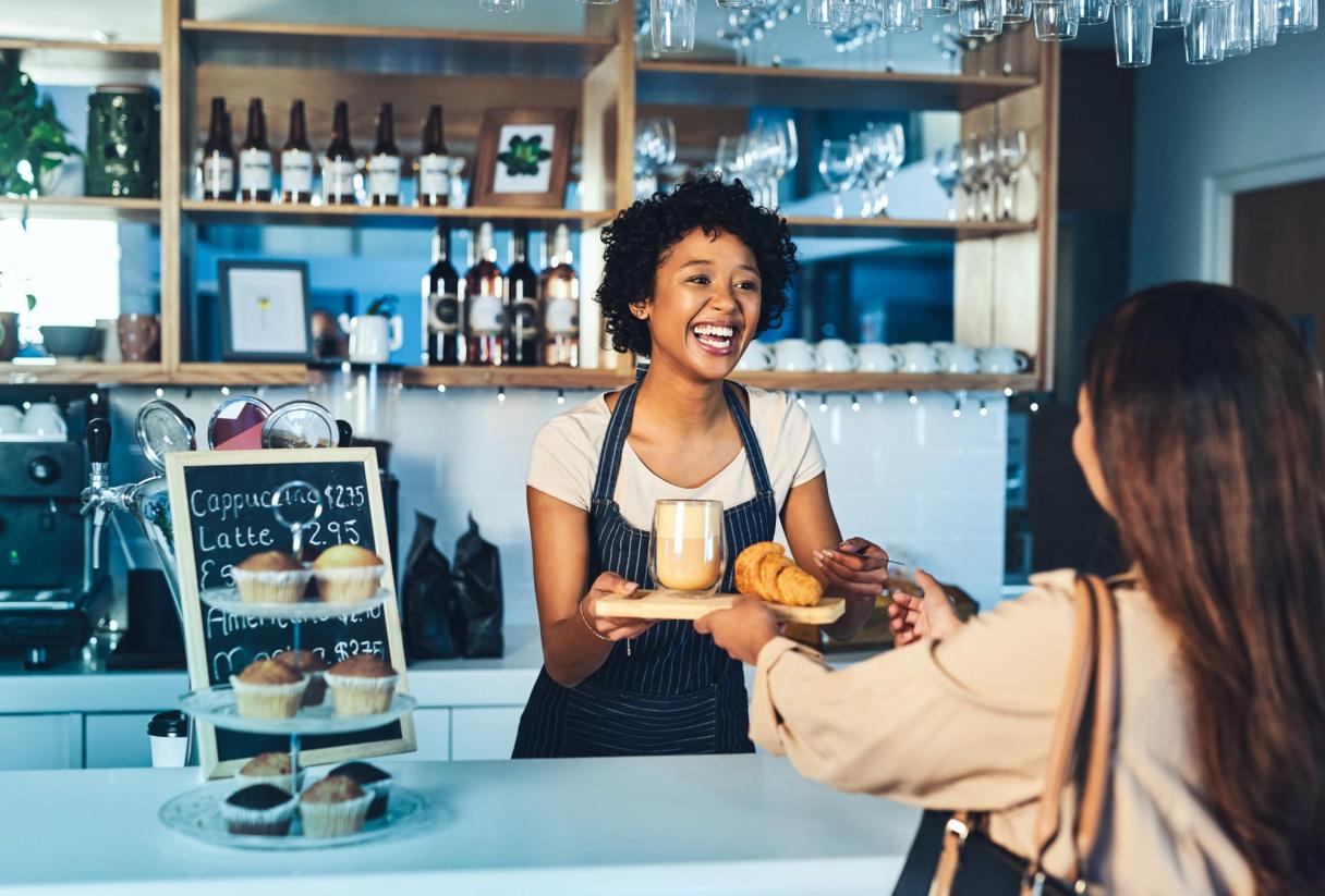 A smiling barista hands a coffee and croissant to a customer at a cafe.