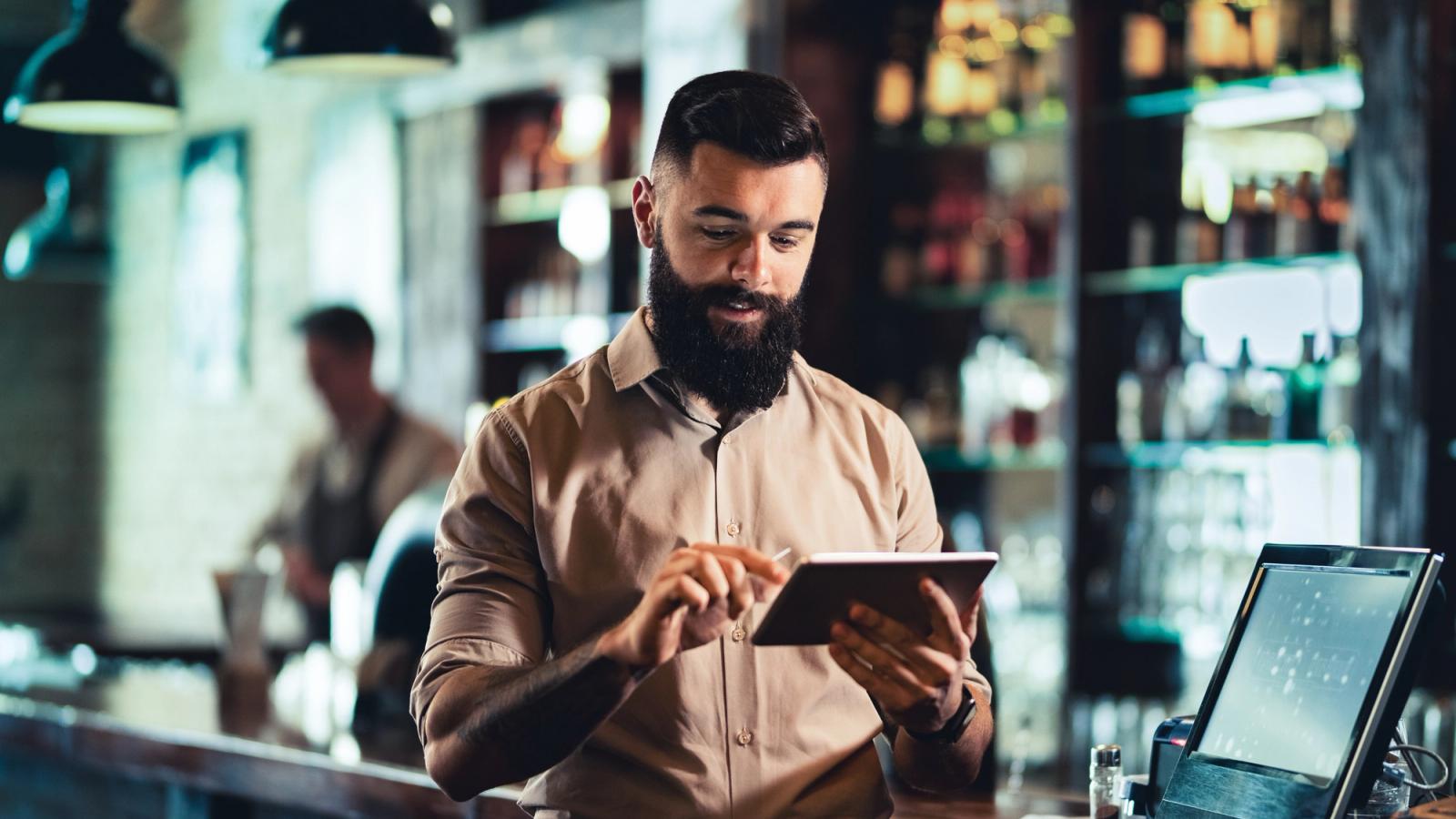 A bearded man uses a tablet at a bar counter, with a blurred second person and a till screen behind