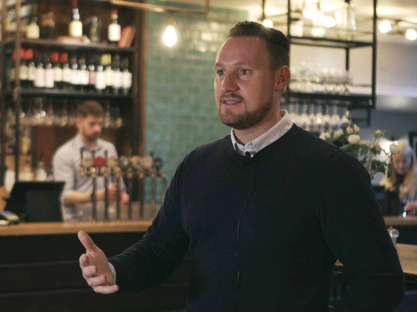 Liam Powell gesturing as he speaks in a pub, with someone at the beer taps behind him