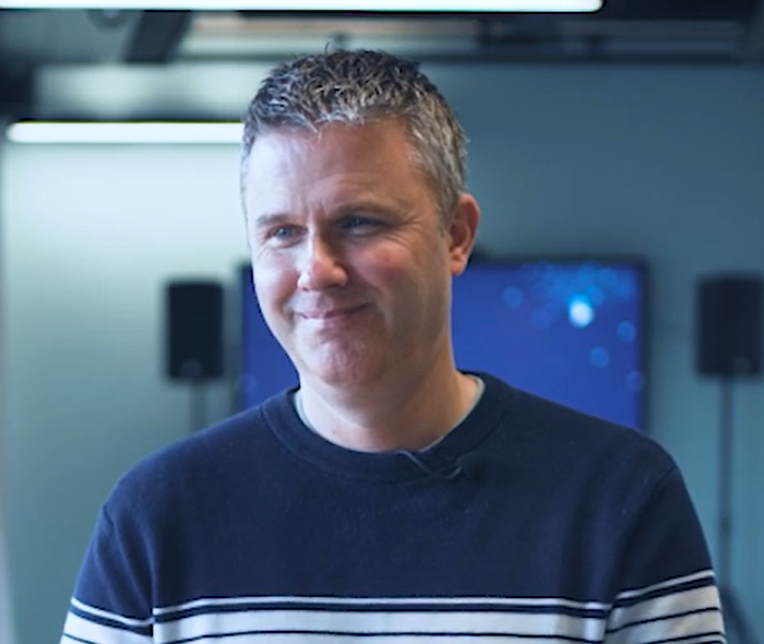 Lee Spooner, smiling in a navy striped jumper in a room with large screens