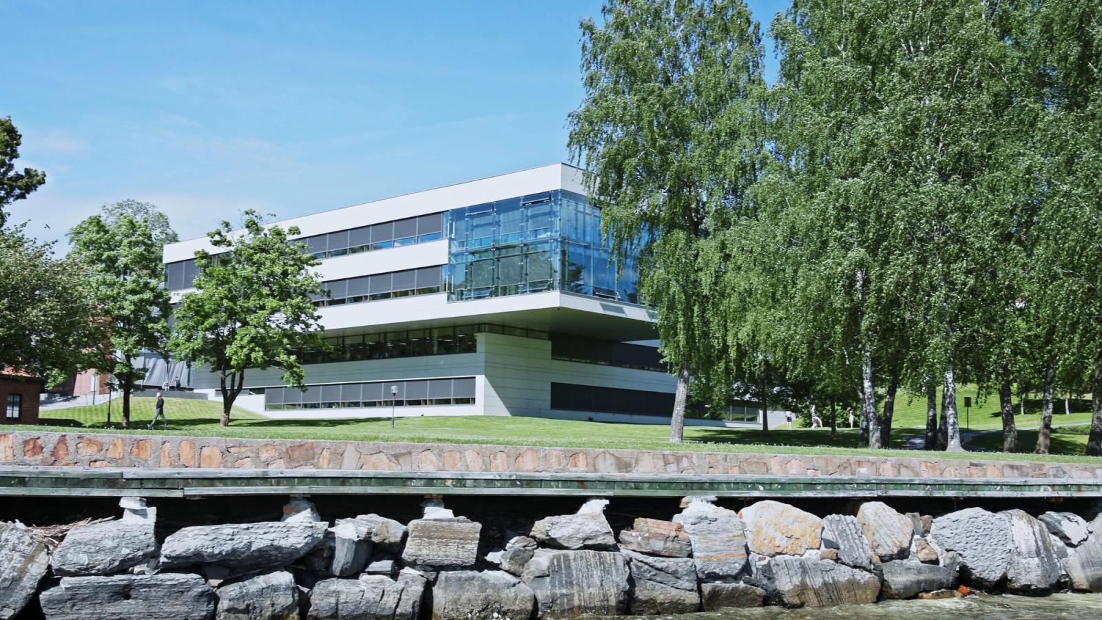 Modern white and glass office building on a lawn behind trees, with a stone shoreline in front