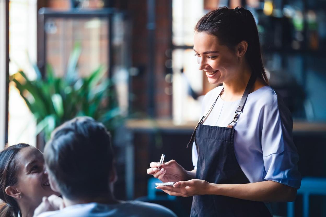 A smiling waitress takes an order from two customers in a restaurant.