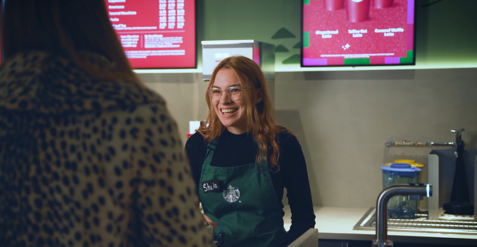 Smiling Starbucks barista in a green apron greeting a customer at the counter
