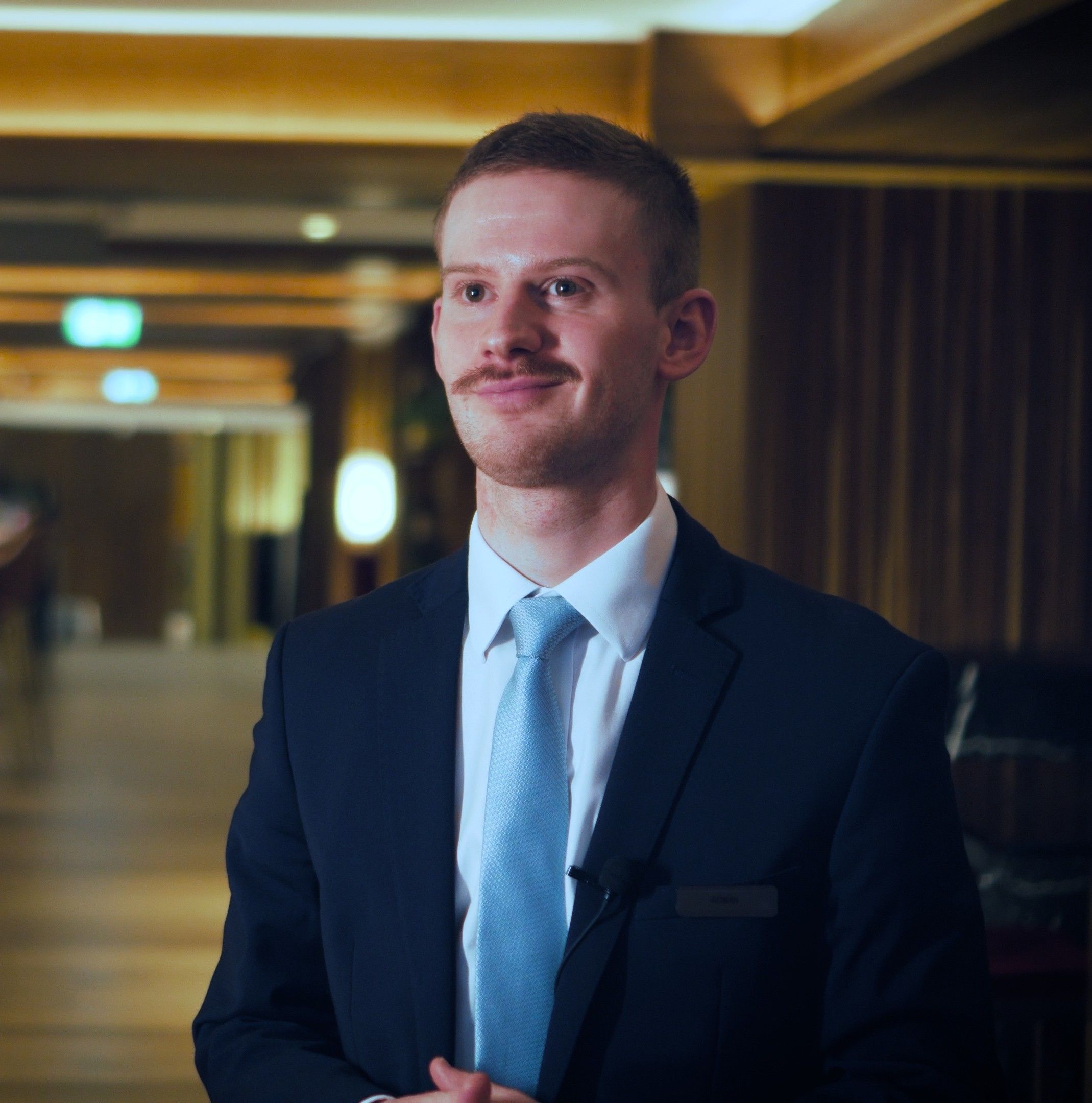Rowan, in a dark suit and light blue tie with a name badge, smiles during a filmed interview