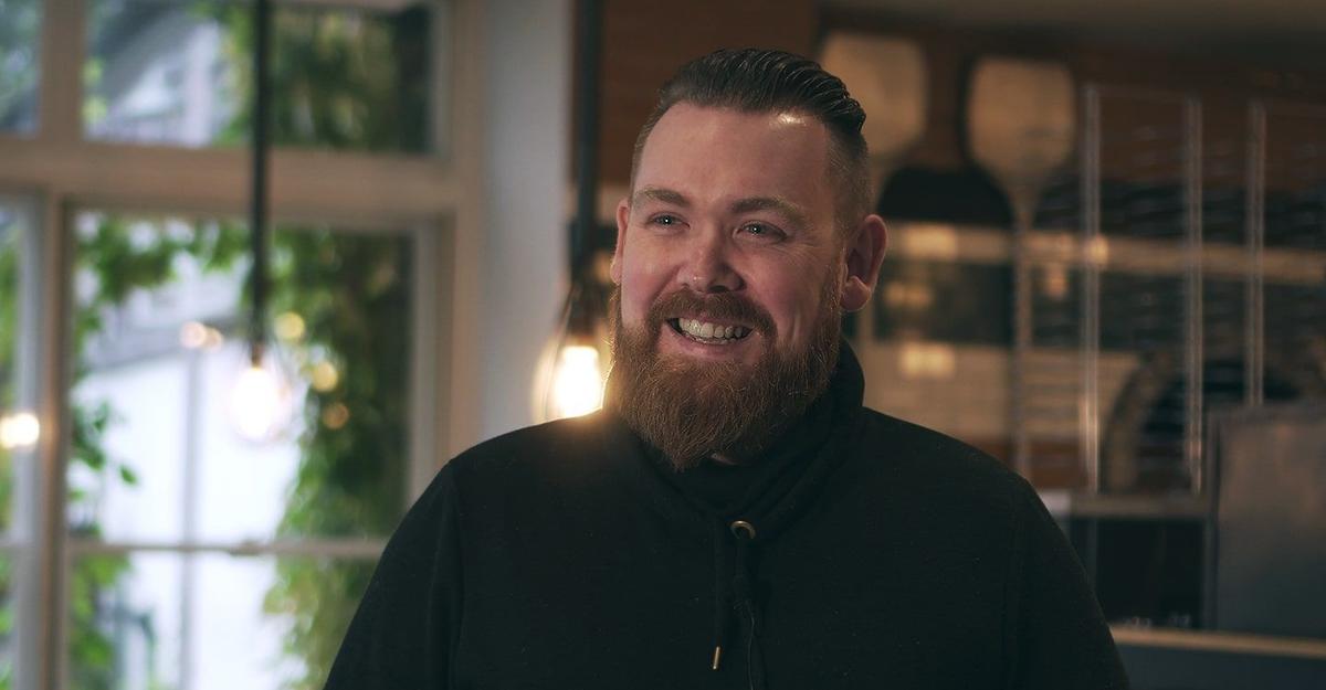 Smiling bearded man in a black shirt in a pub with hanging lights and a buttoned leather bench