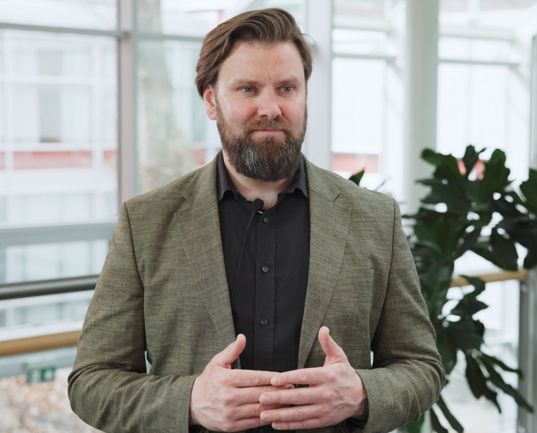 Bjørnar Krukhaug, bearded man in a blazer speaking indoors by a glass wall