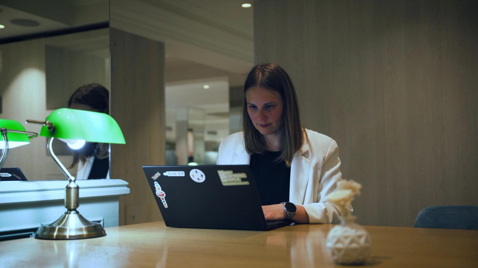 A woman in a white blazer works on a sticker-covered laptop at a wooden table beside a green desk lamp