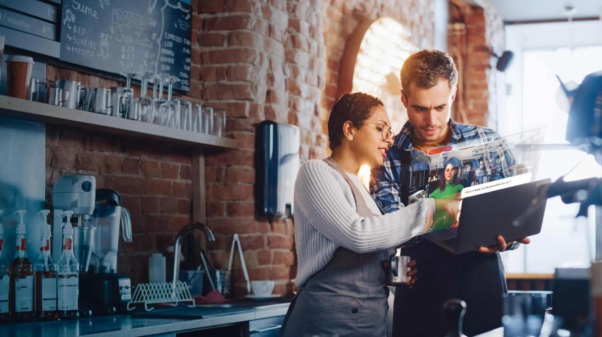Two people in a cafe looking at a laptop displaying a holographic interface.